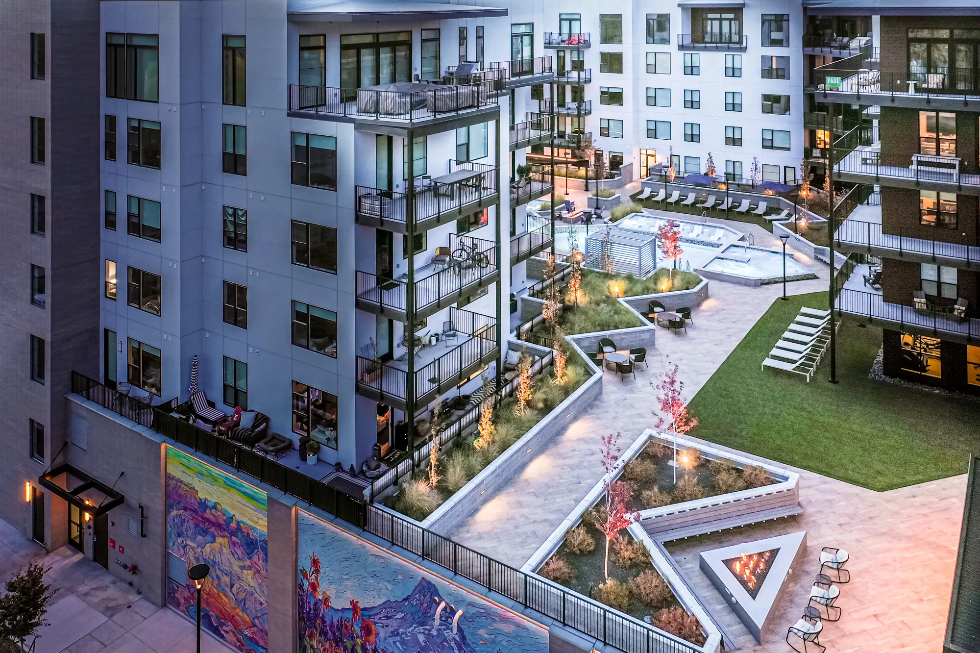 The Grandeur at dusk seen from above, chevron lawn terraces and lit murals stepping down to the pavilion, warm windows across the courtyard