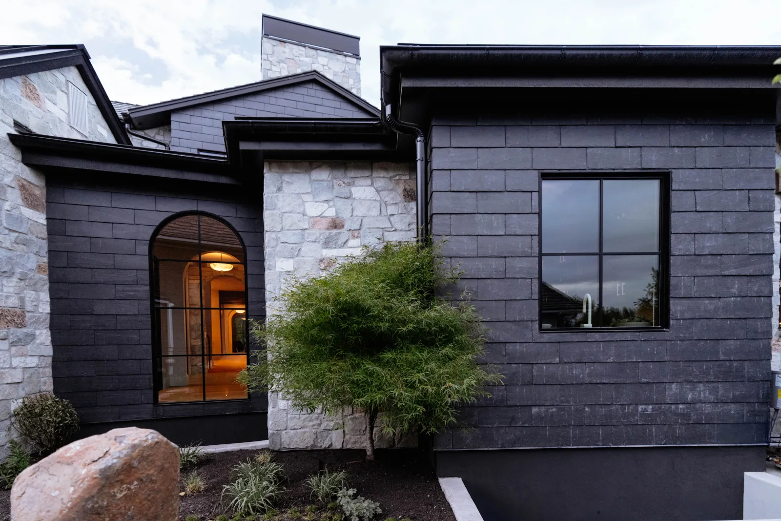 A specimen Japanese maple against the dark siding and stone, an arched window lit beside it