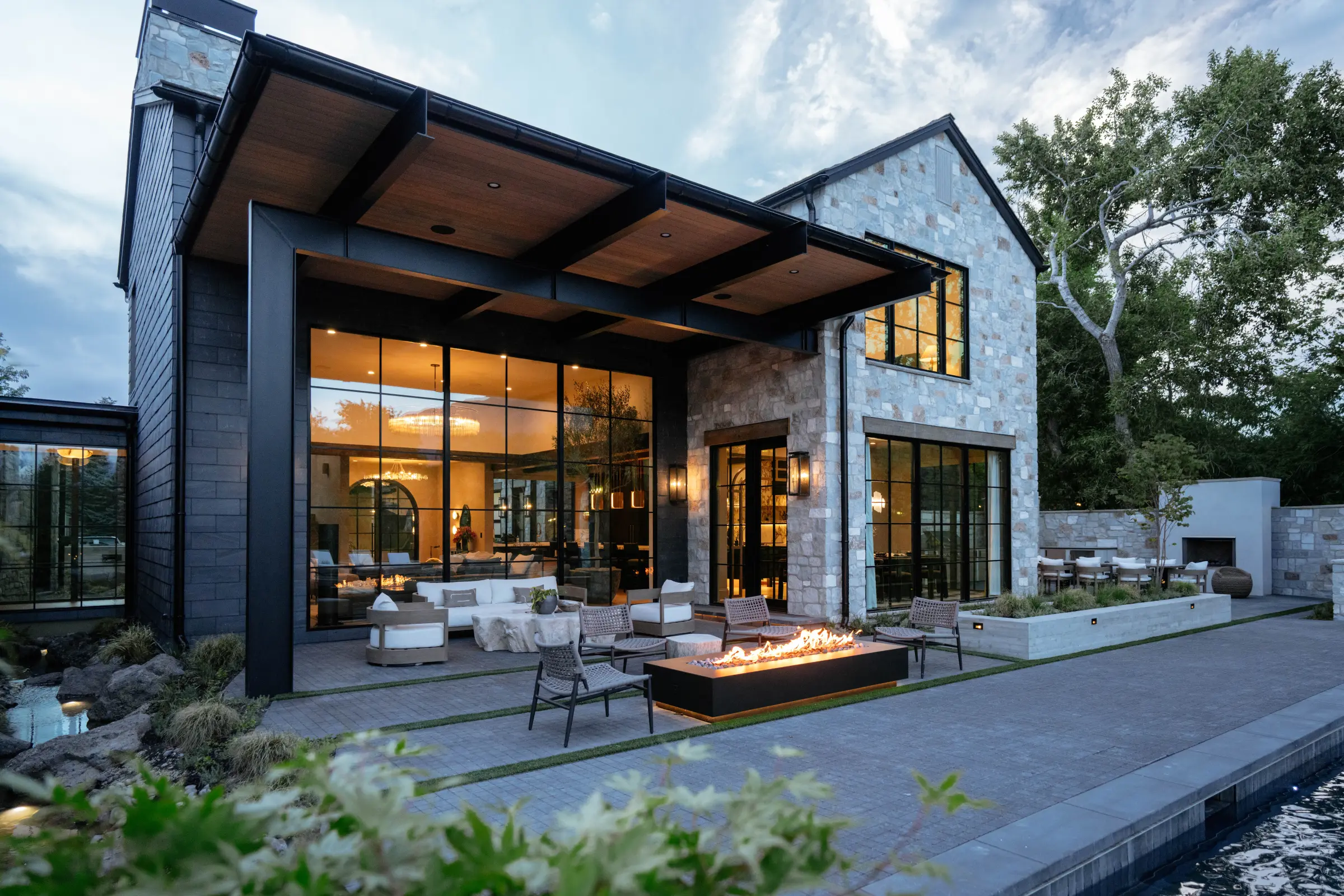 Hidden Oak Haus at dusk, a lit fire table under the steel pergola with the glass wall of the house beyond