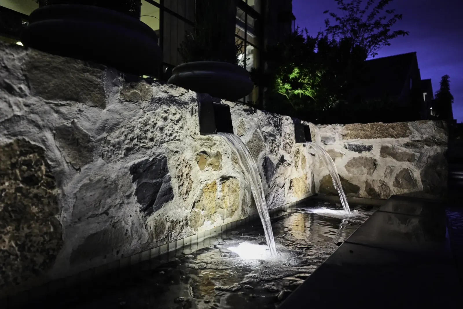 Modern Elm at night: water scuppers pouring from the lit stone wall into the pool