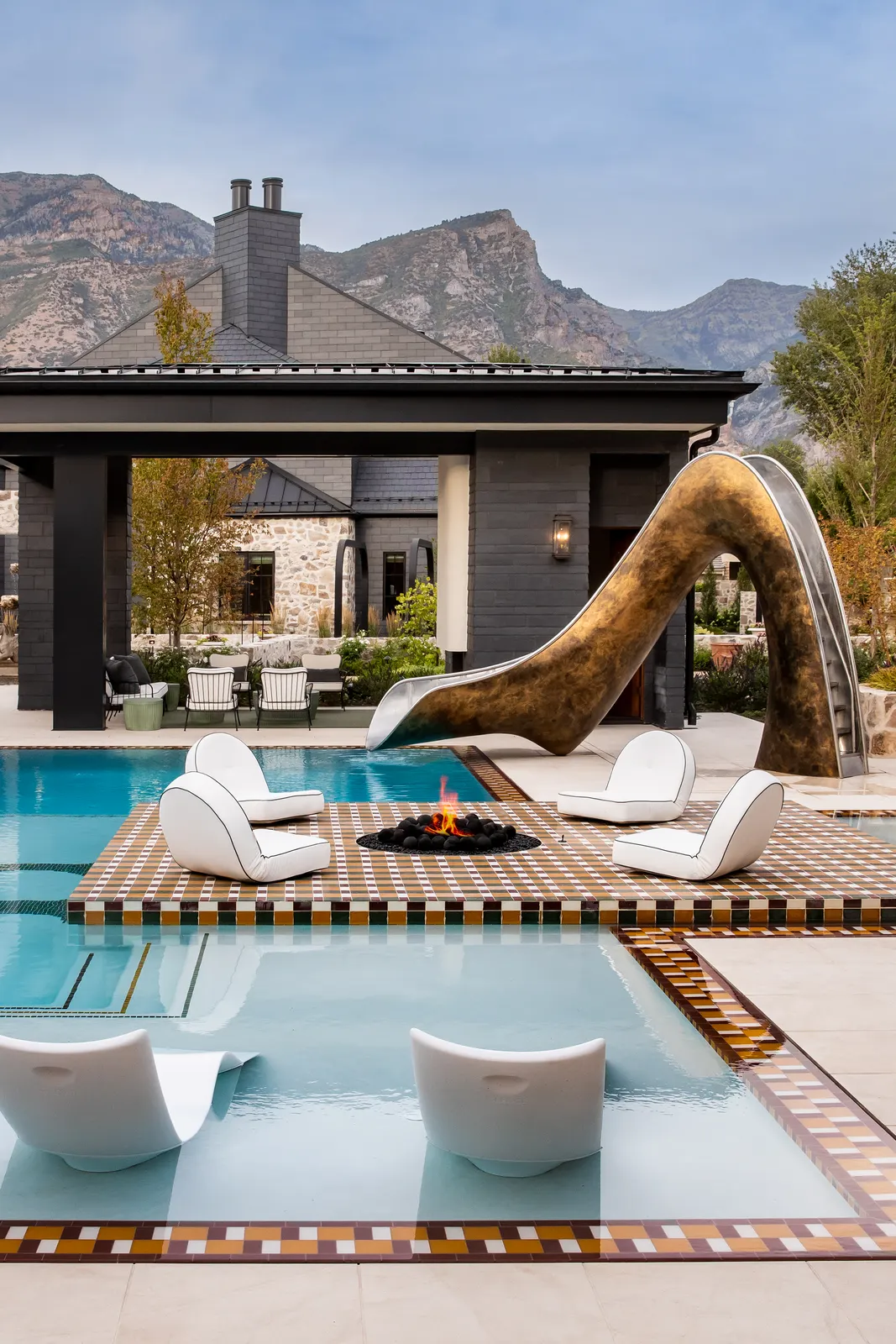The Modern Elm pool: a bronze slide curving into the water, a fire lit between sculpted white loungers, and mountains rising behind the pavilion