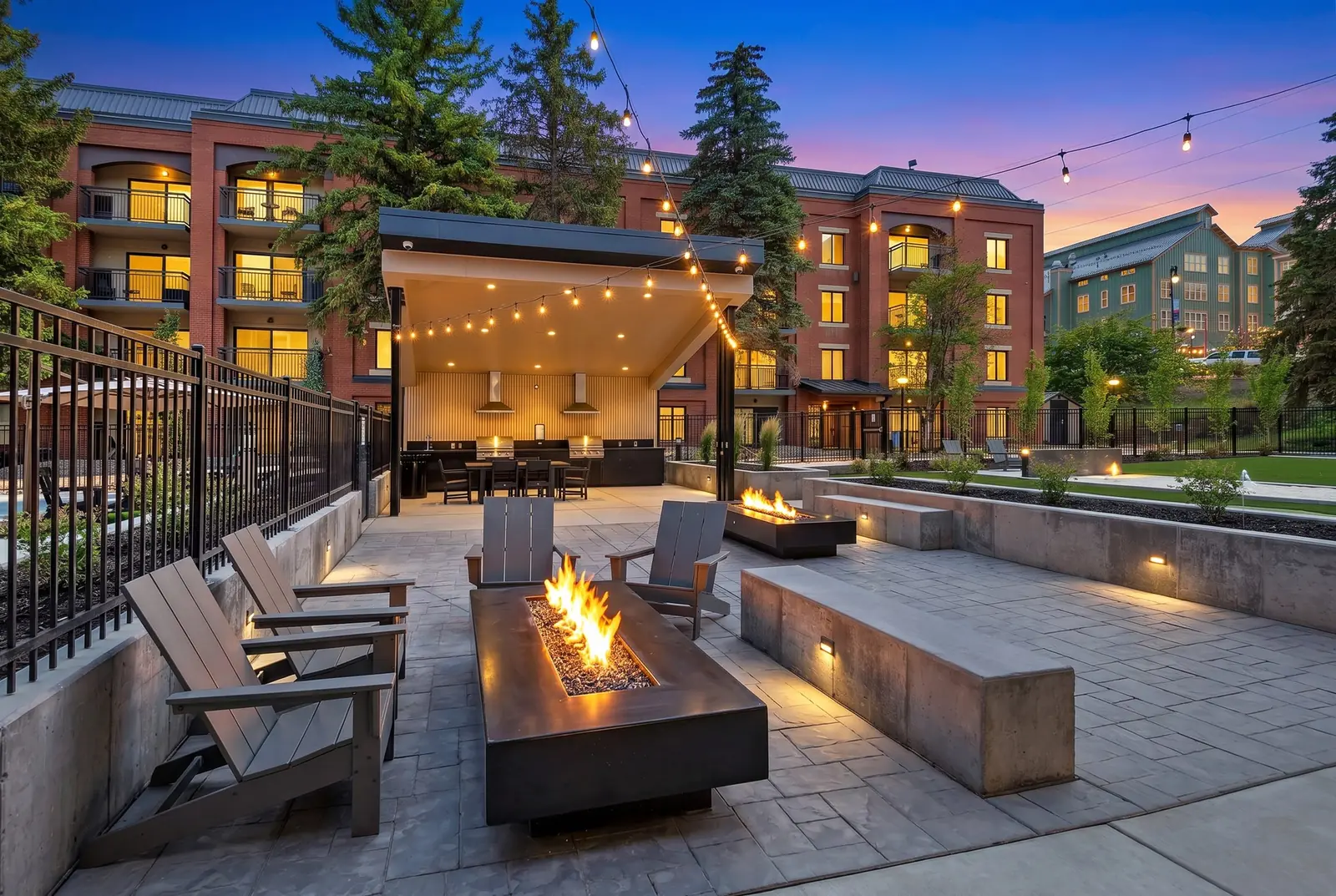 Fire tables and Adirondack chairs in the lounge at dusk, string lights overhead and the covered outdoor kitchen behind
