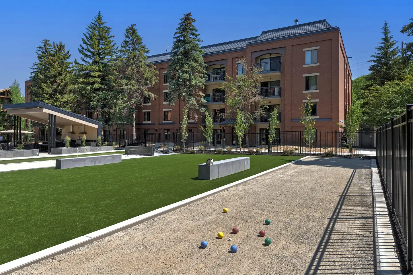 Bocce court beside the lawn with the balls out on the court, a condo building and tall evergreens behind