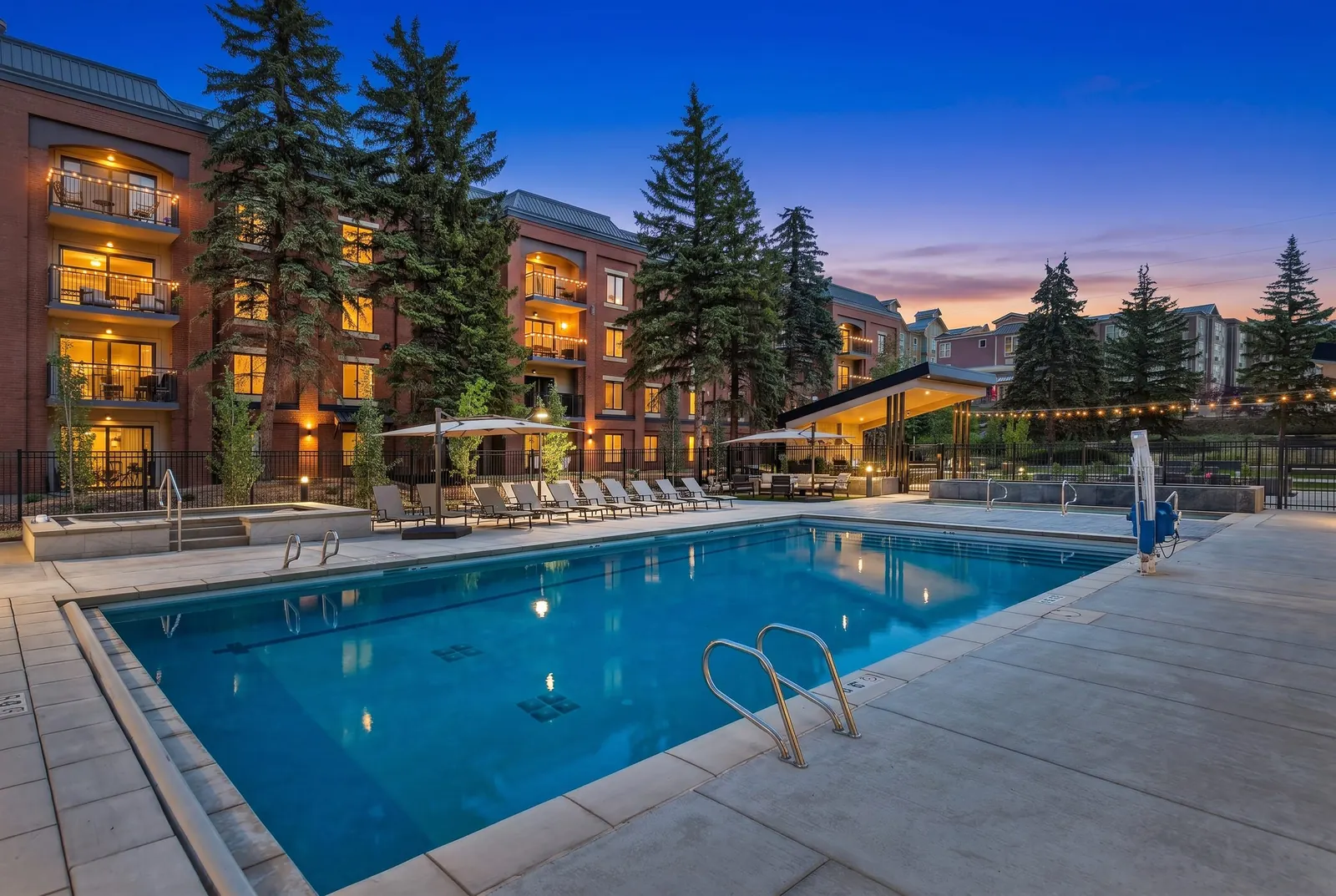 The pool at dusk, lounge chairs and umbrellas along the far side and the condo buildings lit up behind