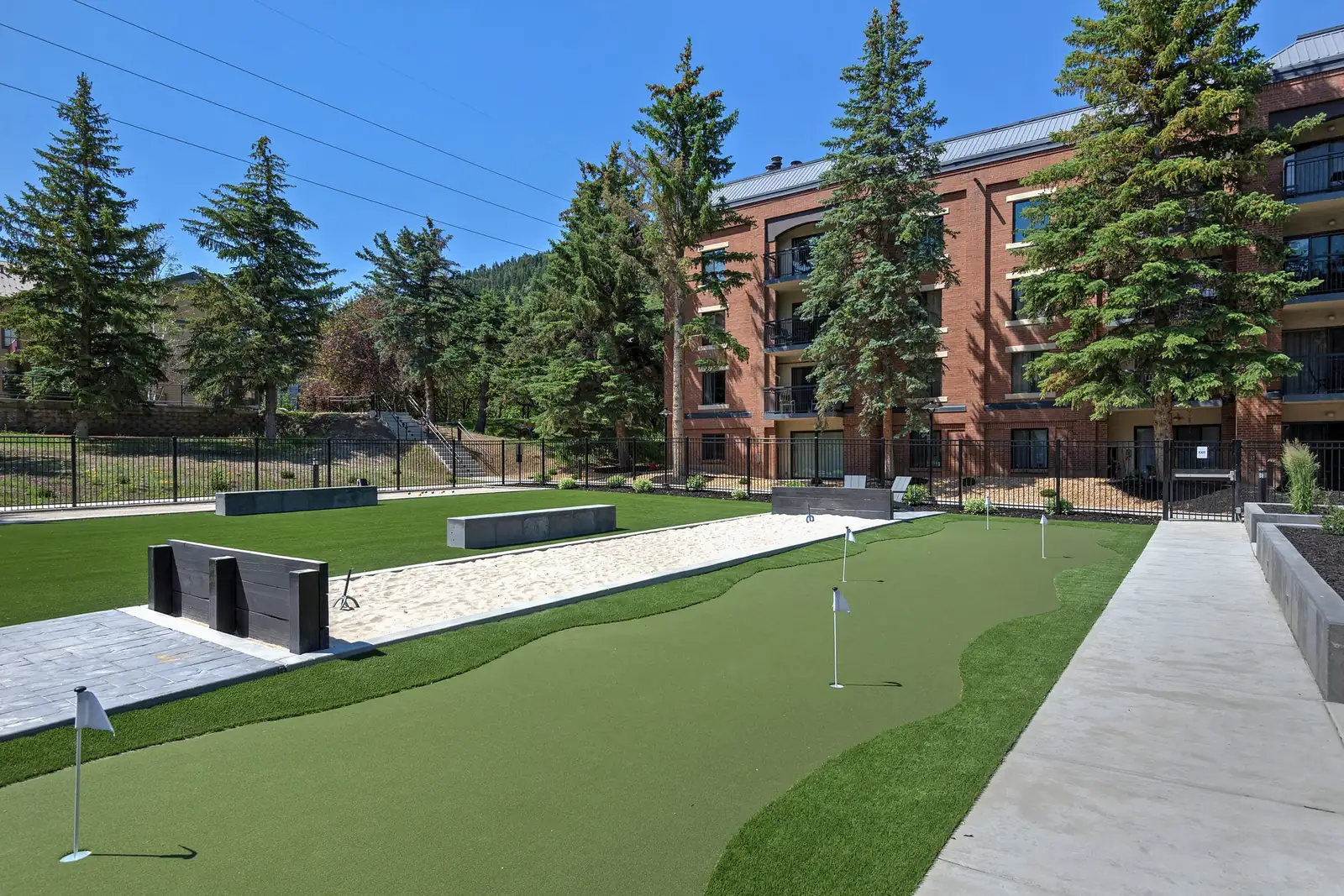 Putting green with flags in front of the bocce court and lawn, evergreens and a condo building behind